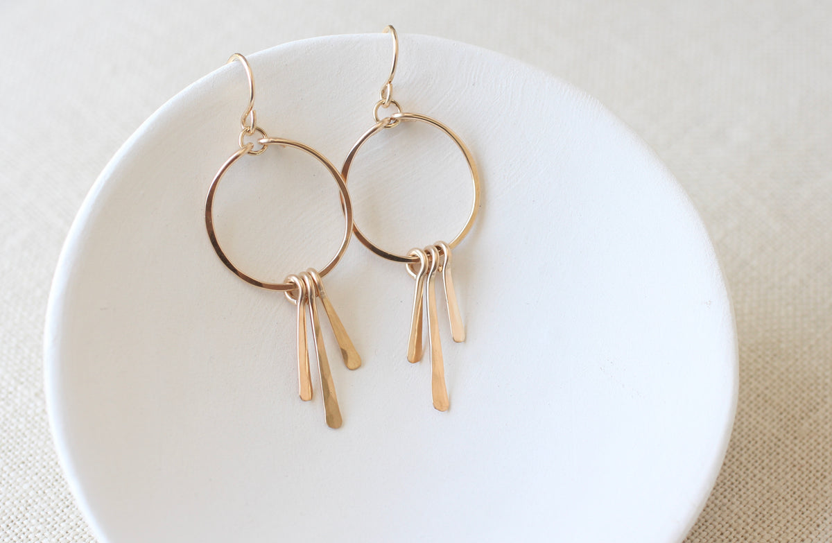 A pair of gold-toned drop hoop earrings with three dangling bar elements, displayed on a white ceramic dish over a beige fabric background.