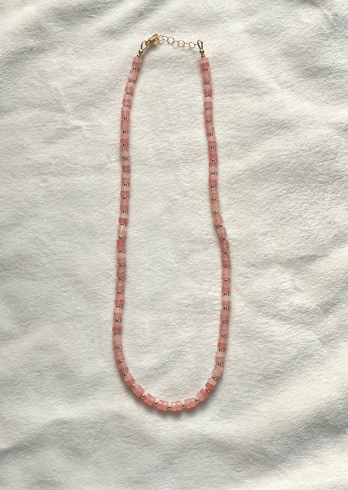 A pink beaded necklace with small gold-toned spacer details and a gold clasp, laid flat on a textured white fabric background.