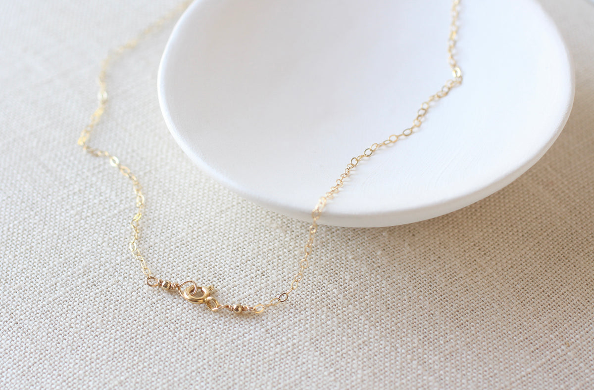 A delicate gold chain necklace with a small bead and heart charm, draped over a white ceramic dish on a textured beige linen surface.