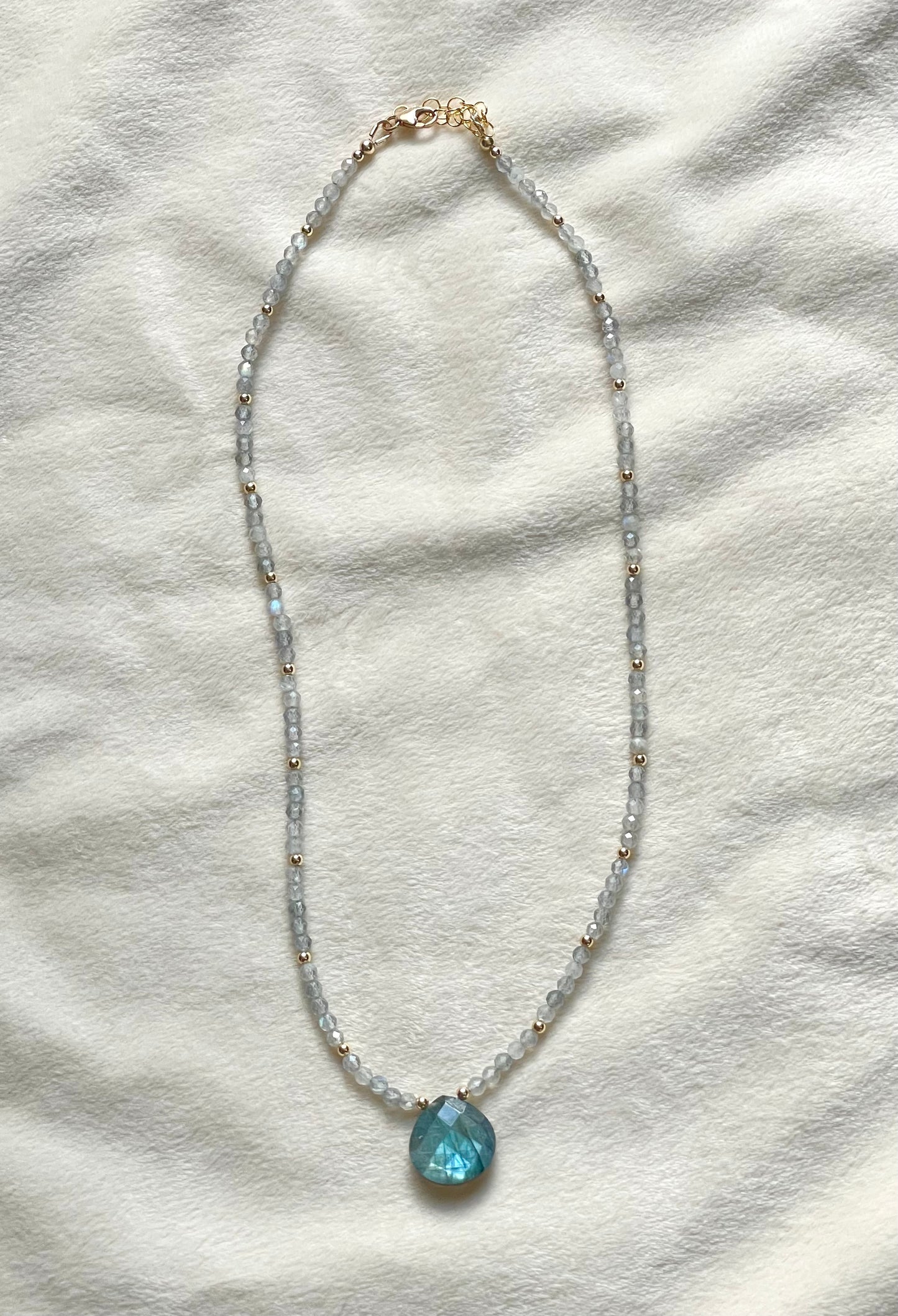 A delicate beaded necklace featuring light blue-gray faceted beads, small gold spacer beads, and a faceted light blue-green pendant, displayed on a textured off-white fabric background.