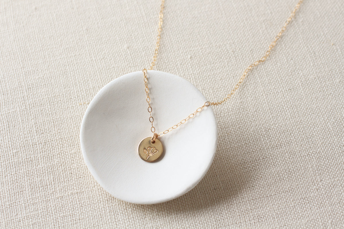 A delicate gold chain necklace with a small round stamped flower gold pendant, displayed on a white ceramic dish over a beige linen background.