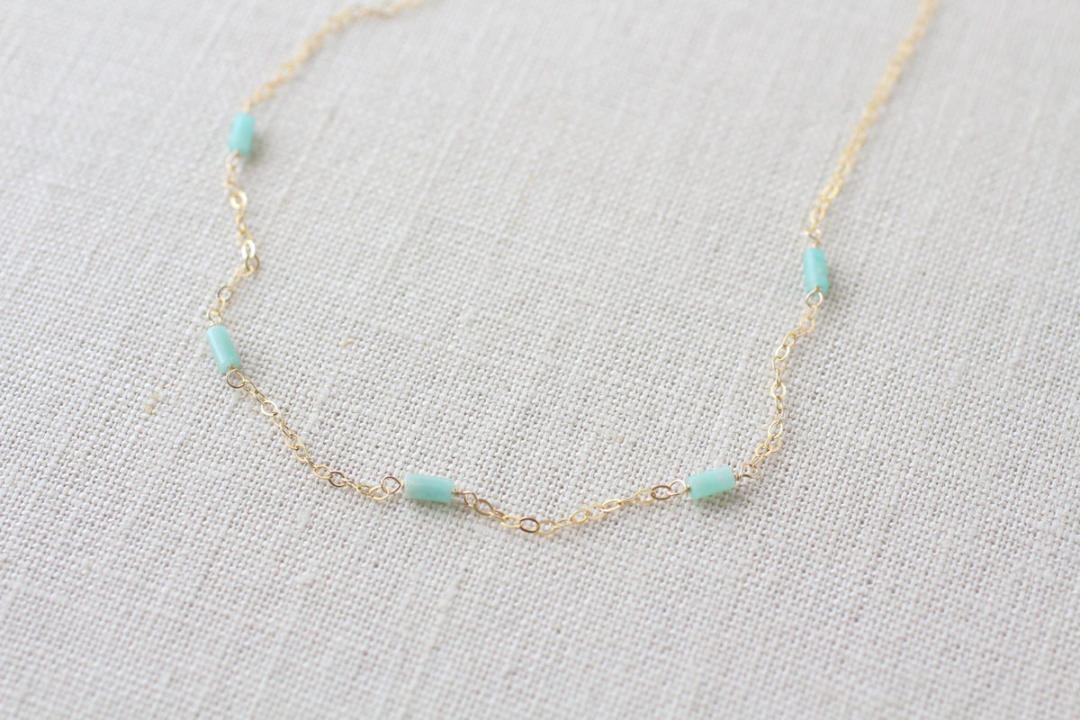 A delicate gold chain necklace featuring tiny, evenly spaced light green colored cylindrical beads, displayed on a textured beige linen fabric background.