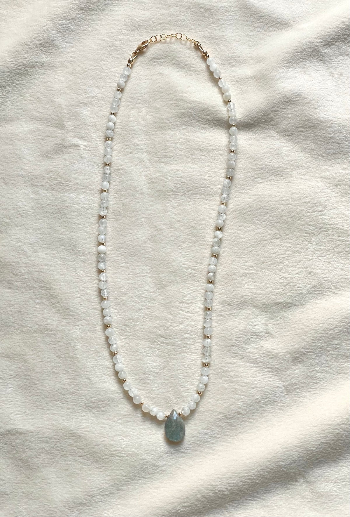 A beaded necklace with small white and gold beads, a gold-toned clasp, and a smooth oval light blue pendant, laid flat on a textured off-white fabric background.