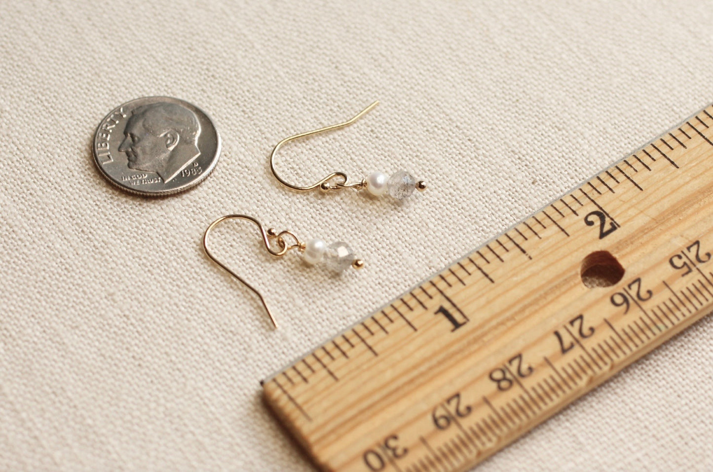 Gold-toned fishhook earrings with small clear pearl drops, shown on a beige fabric surface
next to a U.S. dime and a ruler