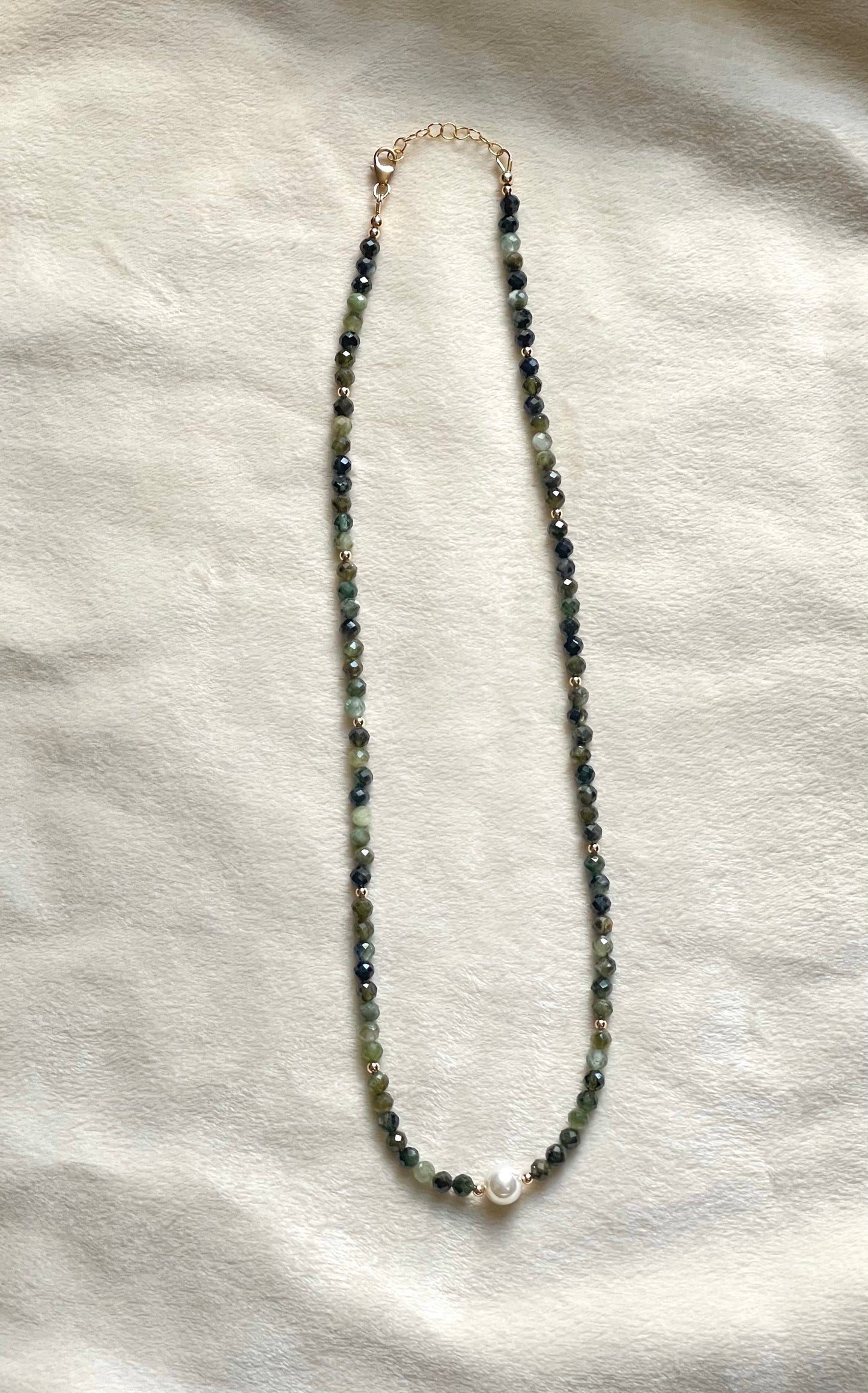 A beaded necklace with small dark green beads, a white pearl accent at the bottom center, and a gold-tone clasp, laid flat on a textured off-white fabric background.