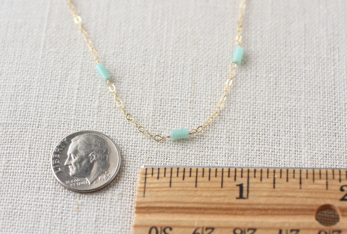 A delicate gold chain necklace with three small mint green cylindrical beads, displayed on a textured beige fabric alongside a US dime for scale and a wooden ruler.