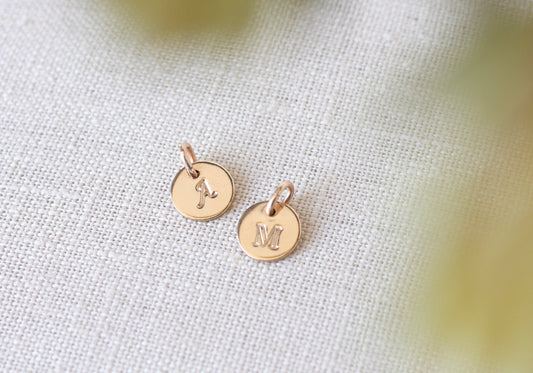 A pair of small round rose gold initial pendants engraved with the letters 'J' and 'M', resting on a textured beige linen fabric background with soft, out-of-focus yellow-green bokeh in the foreground.