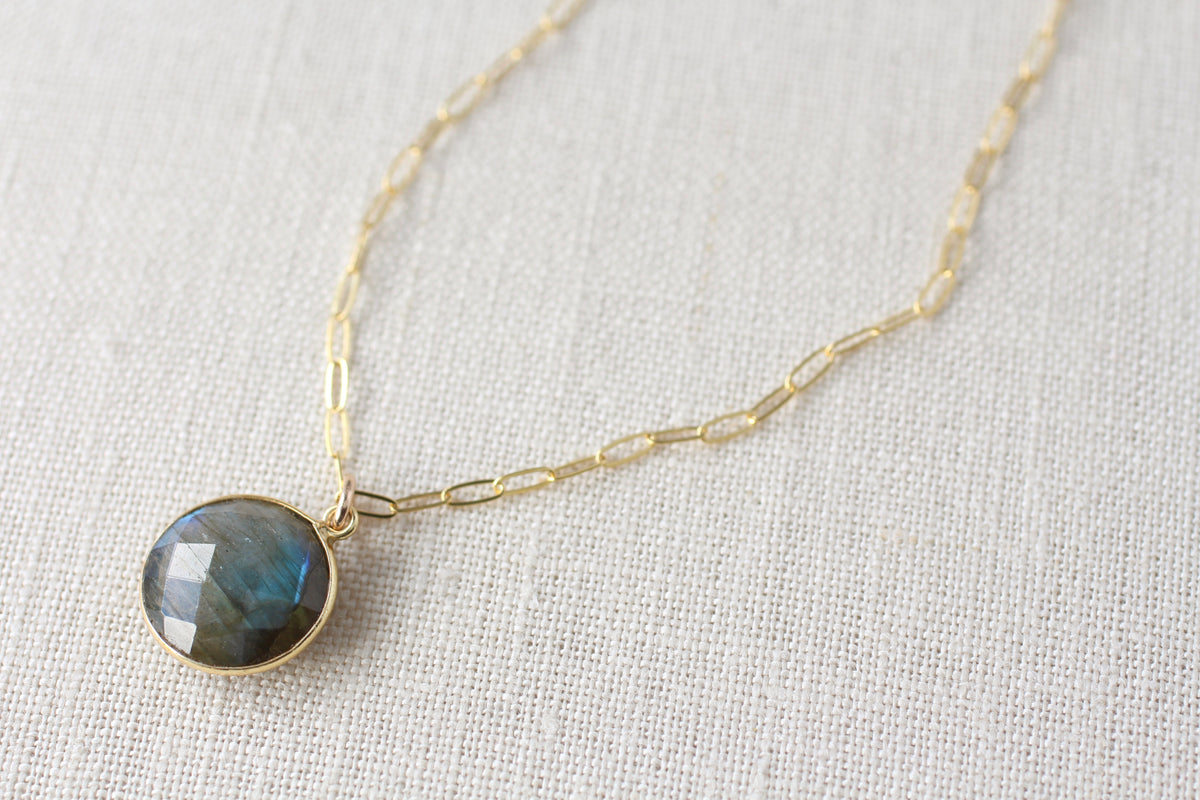 A gold chain necklace with a round bezel-set labradorite pendant, displayed on a textured off-white linen background.