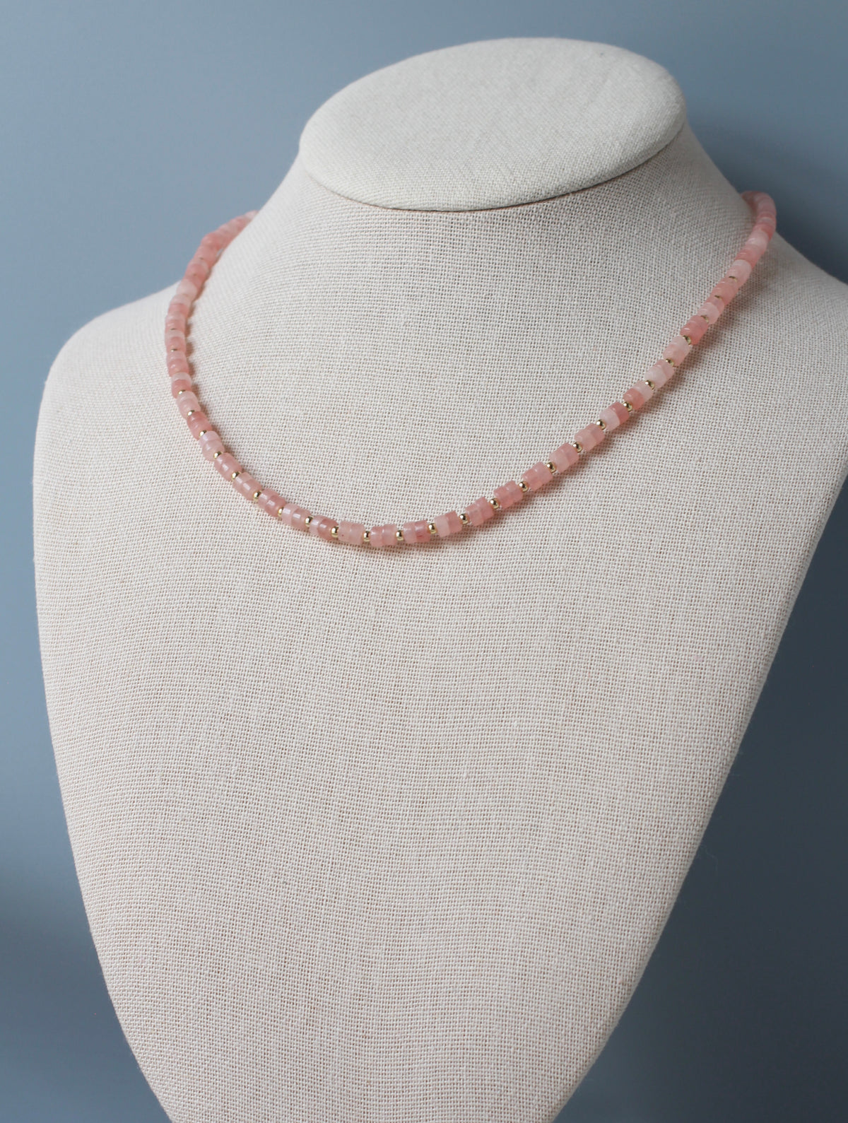 A pink beaded necklace displayed on a cream fabric-covered jewelry bust against a soft blue-gray background.