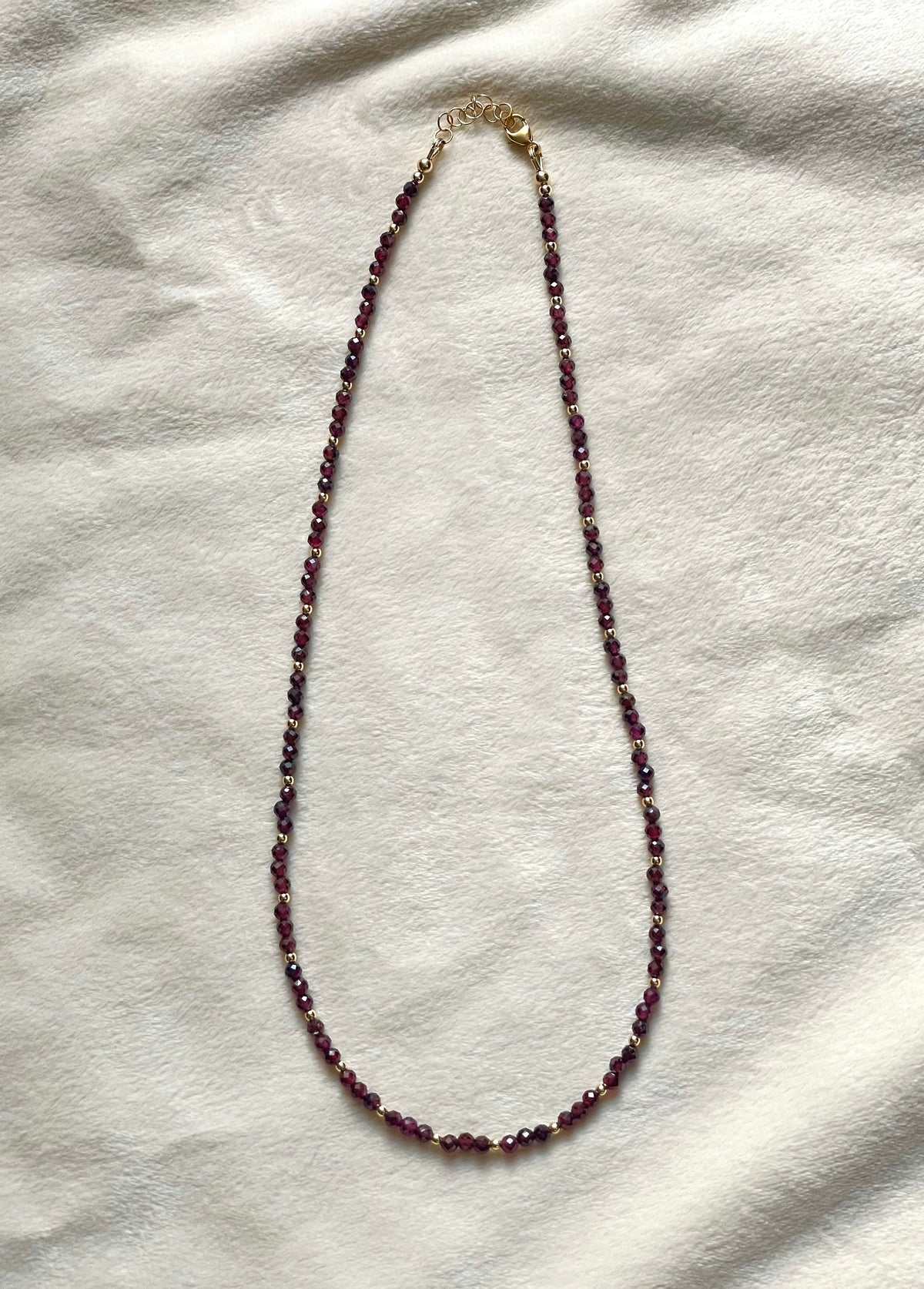 A beaded necklace with small dark red beads separated by tiny gold spacer beads, featuring a gold clasp, laid flat on a textured off-white fabric background.