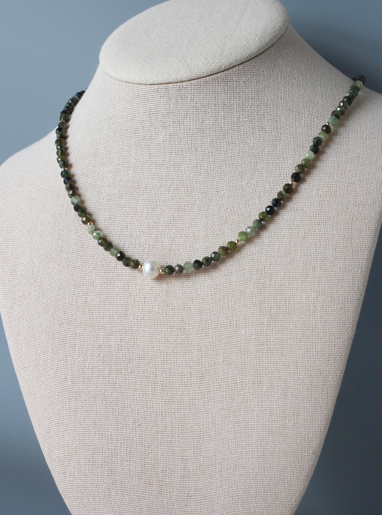 A beaded necklace displayed on a beige fabric jewelry bust. The necklace consists of small dark green and light green beads with a single white pearl bead at the center front.