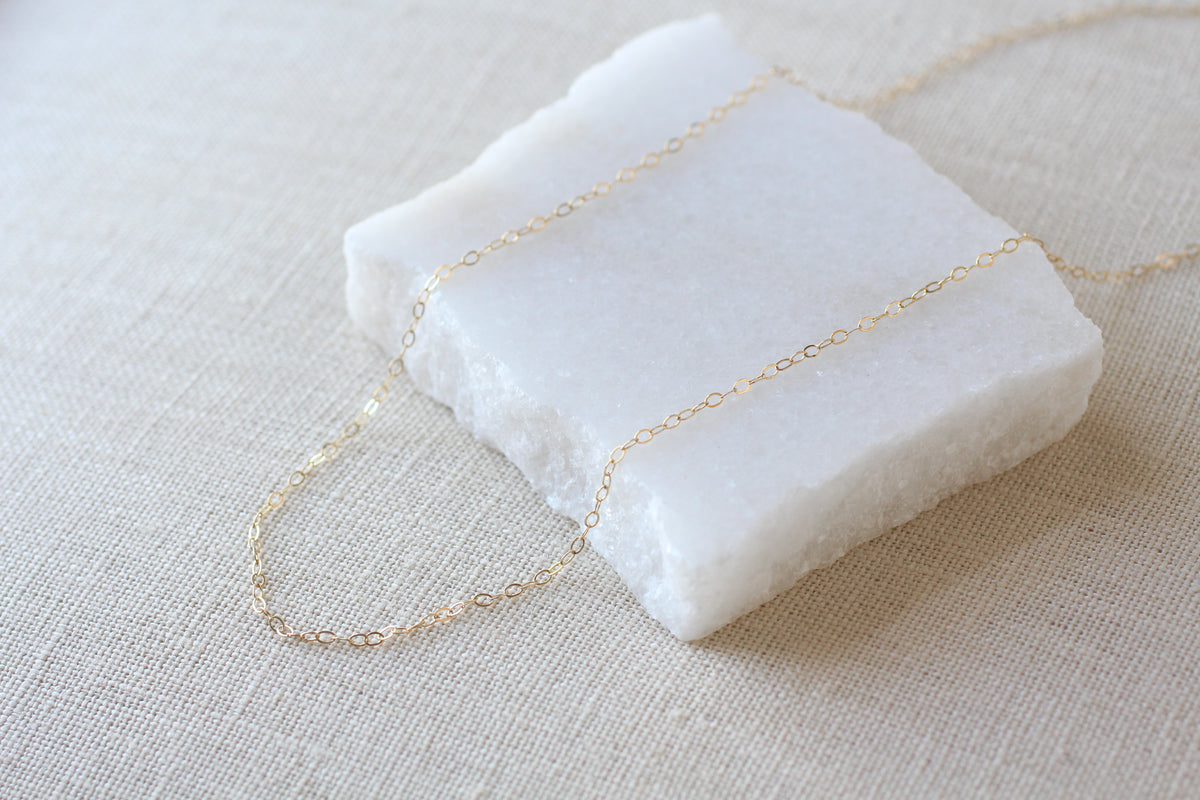 A delicate gold chain necklace draped over a white textured stone block, set against a neutral beige linen fabric background.