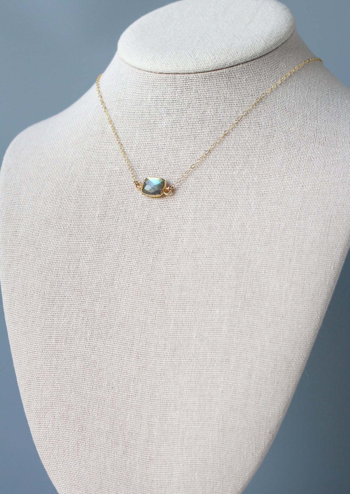 A delicate gold chain necklace with a small organic shaped rectangle stone, displayed on a textured cream fabric jewelry bust against a soft blue-gray background.
