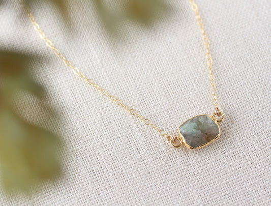 A delicate gold chain necklace with a small, cushion-cut labradorite stone pendant in a gold bezel setting, resting on a textured off-white fabric with soft green leaves blurred in the foreground.