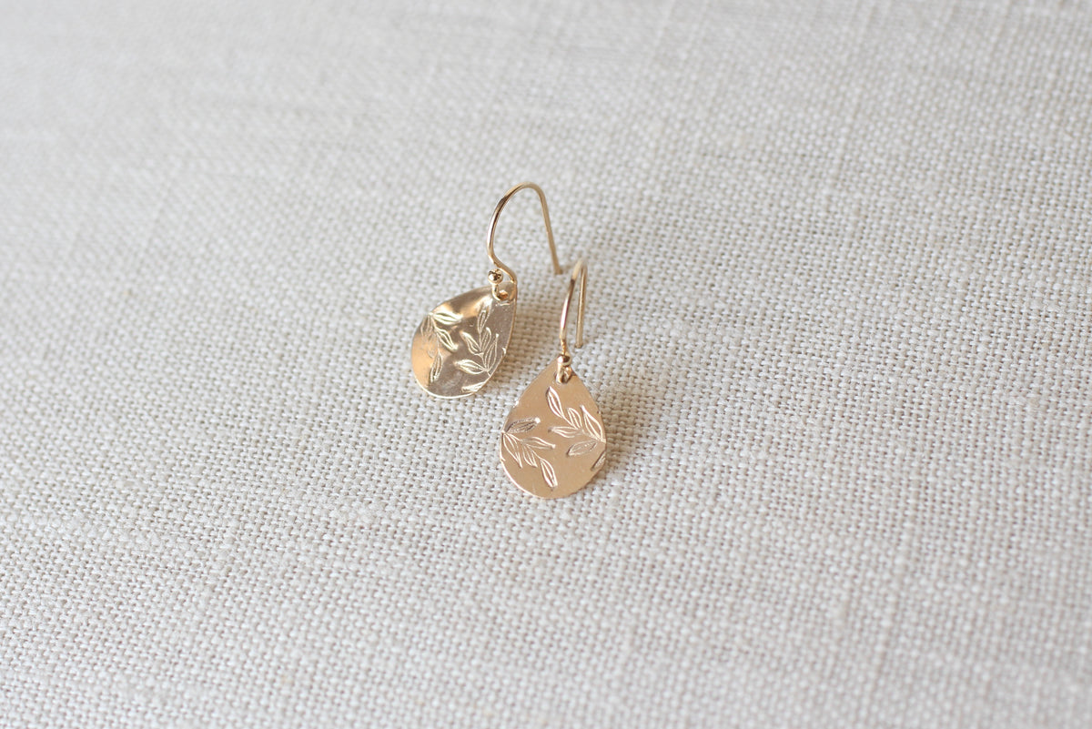 A pair of gold teardrop dangle earrings with a hammered texture, displayed on a neutral beige linen fabric background.