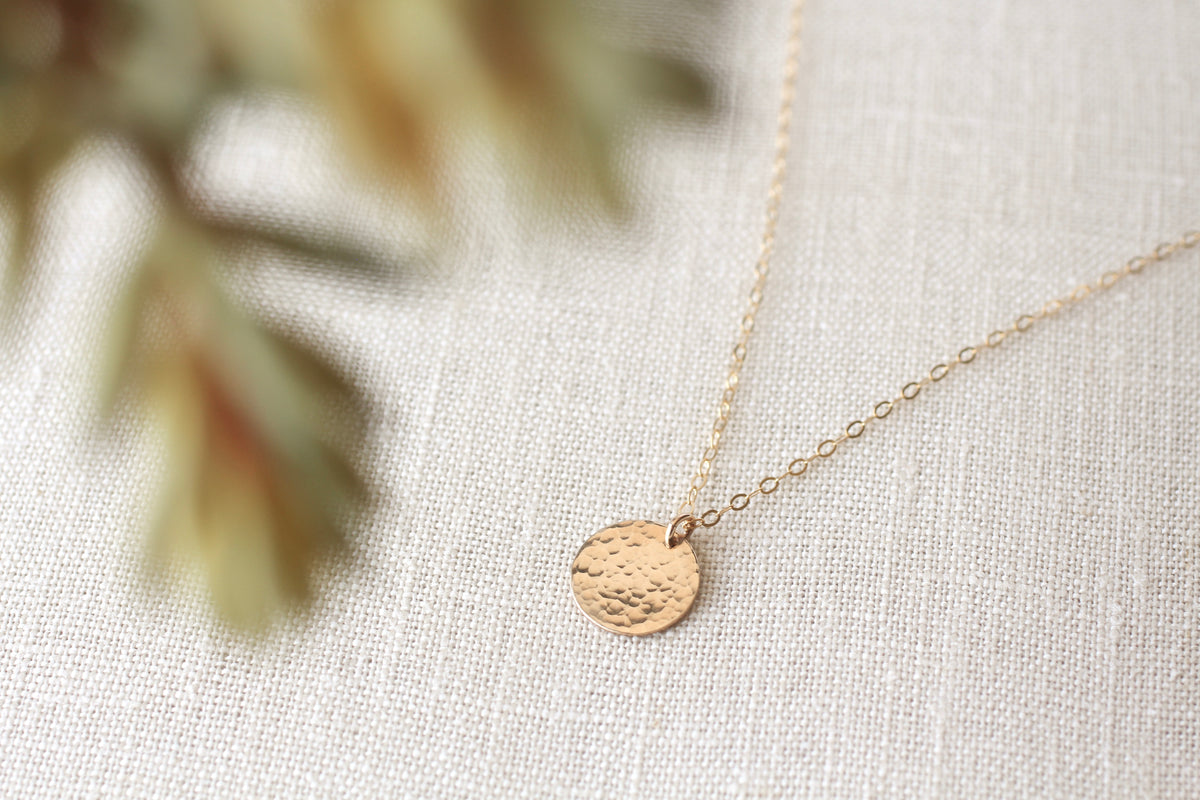 A delicate gold chain necklace with a small, textured circular disc pendant, displayed on a neutral beige linen fabric background with soft, blurred green leaves in the foreground.