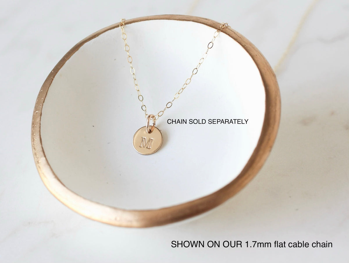 A gold initial pendant necklace with a letter 'N' on a delicate cable chain, displayed on a white ceramic dish with a gold rim. Text overlays state 'CHAIN SOLD SEPARATELY' and 'SHOWN ON OUR 1.7mm flat oval chain’