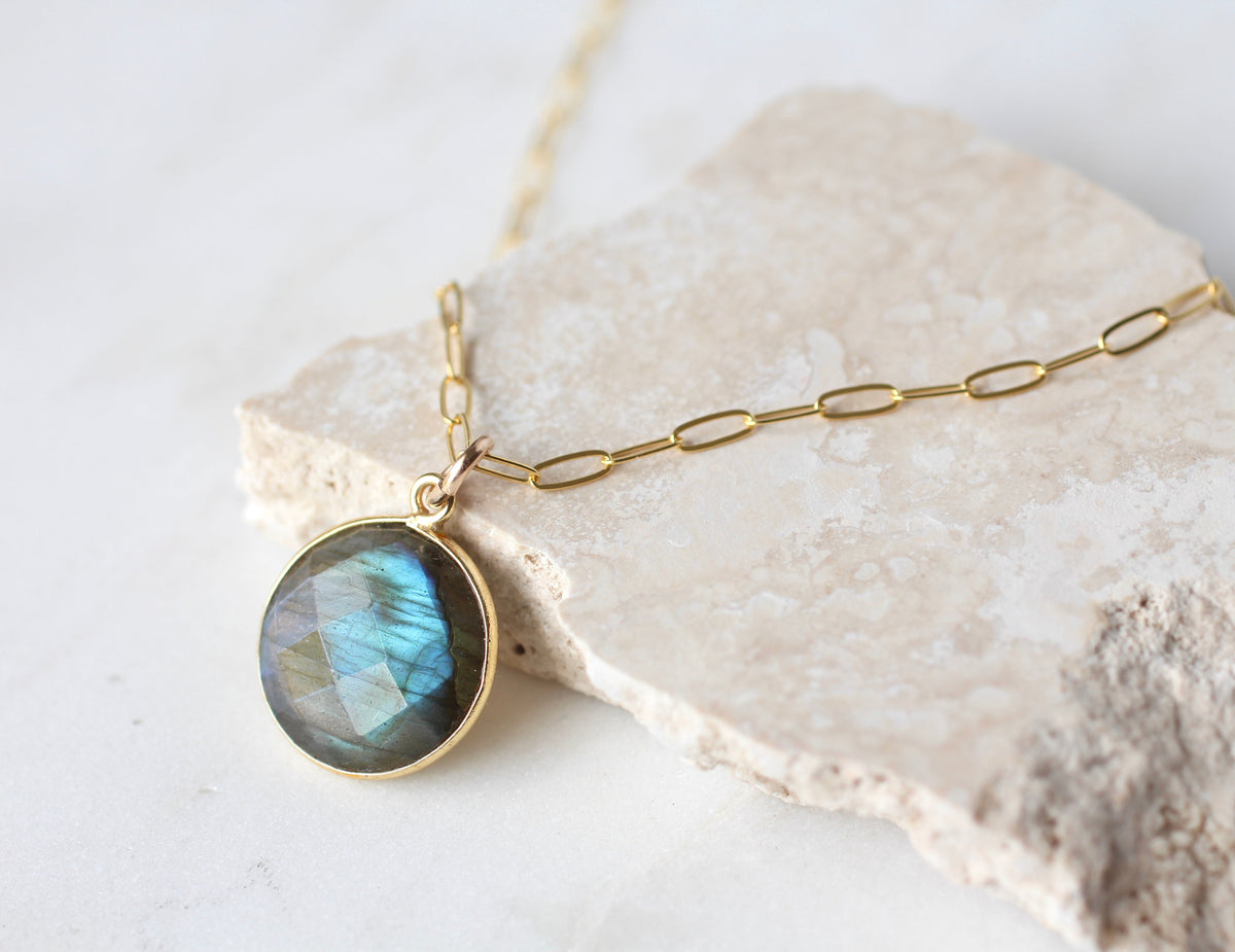A gold chain necklace with a round bezel-set labradorite pendant, displayed on a textured beige stone slab against a light background.