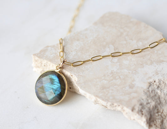 A gold chain necklace with a round bezel-set labradorite pendant, displayed on a textured beige stone slab against a light background.