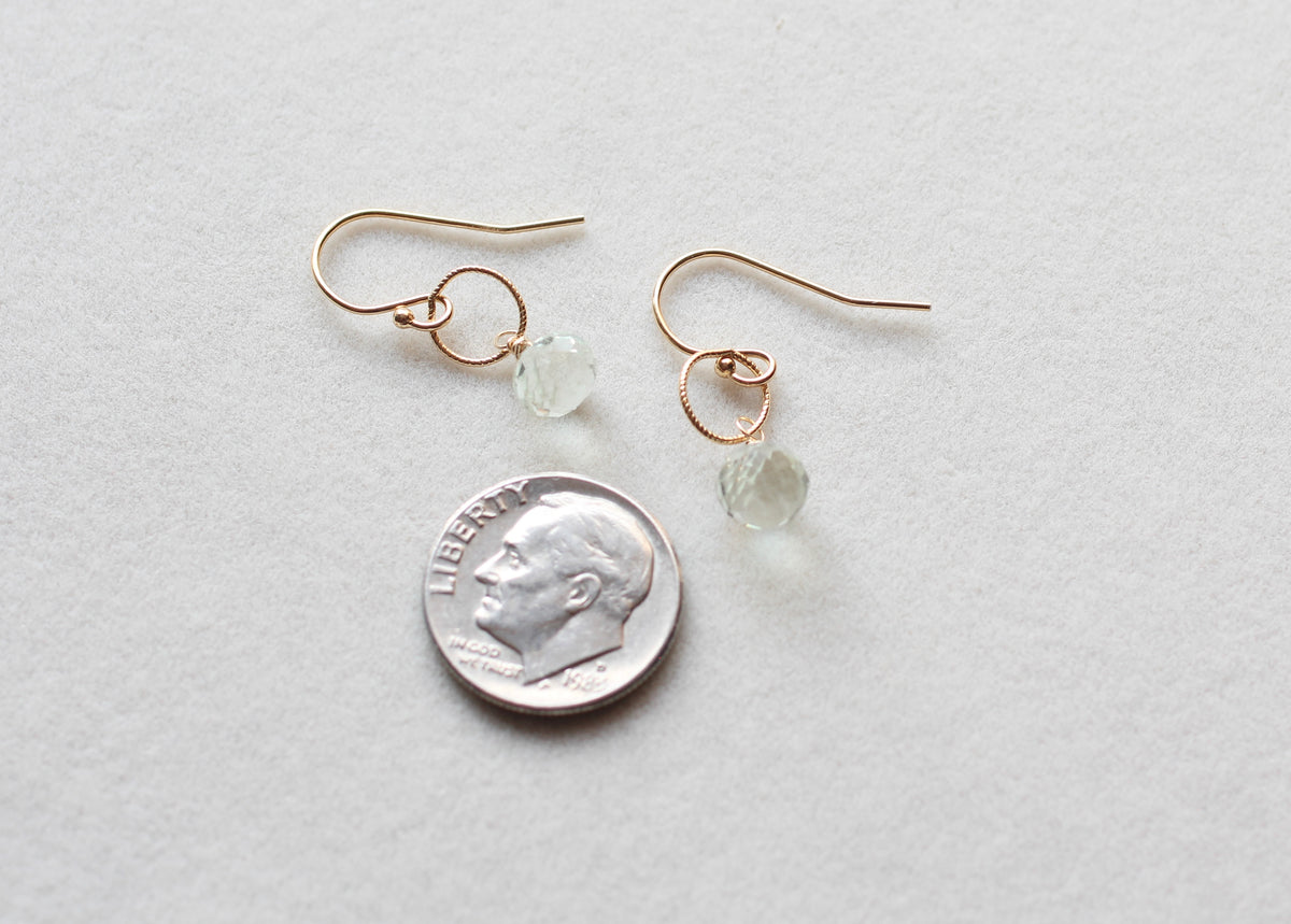 A pair of delicate rose gold wire-wrapped drop earrings with small faceted light green beads, shown next to a US dime for scale on a textured light gray background.