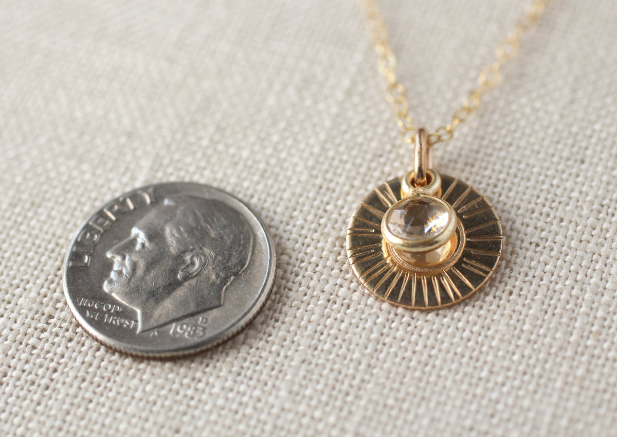 A gold necklace with a circular sunburst pendant and a clear faceted gemstone, shown next to a US dime for scale on a textured beige linen background.