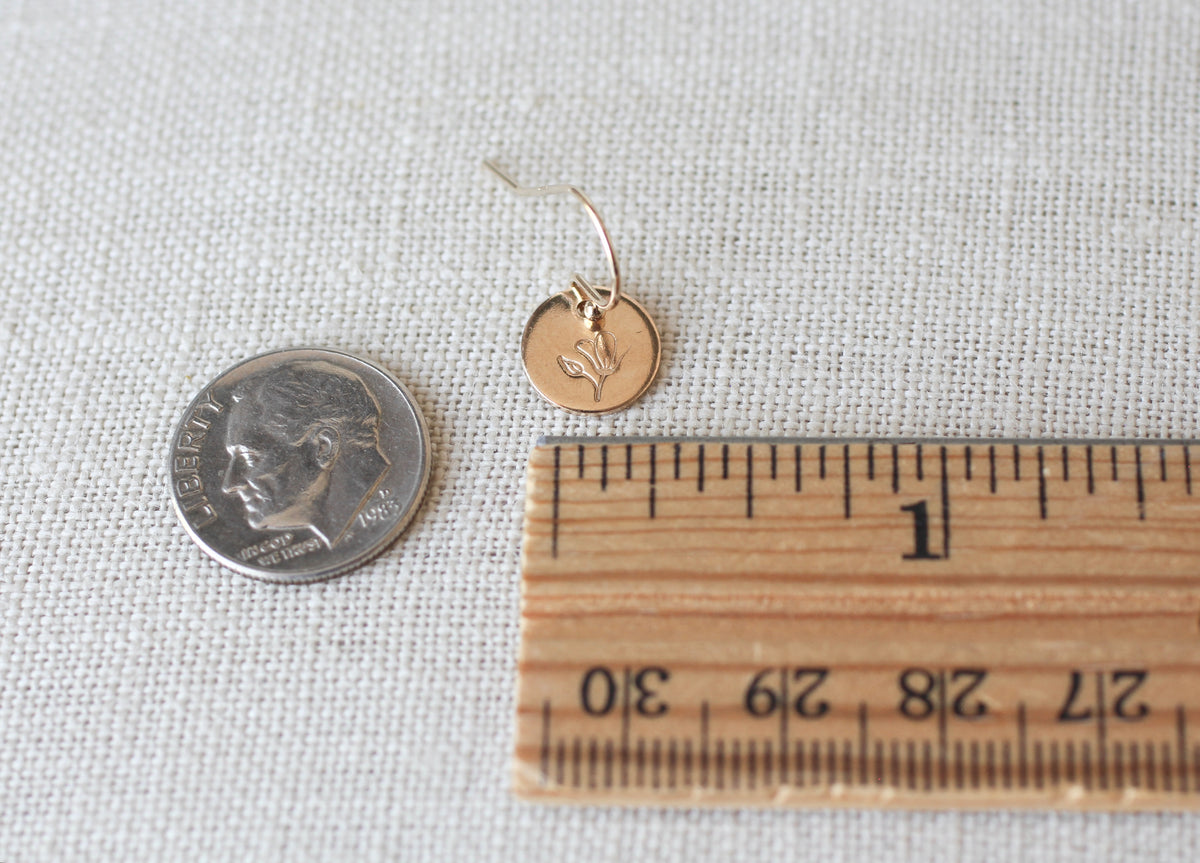 A small gold disc earring with a bird engraving, shown next to a US dime coin and a wooden ruler on a textured beige fabric background.