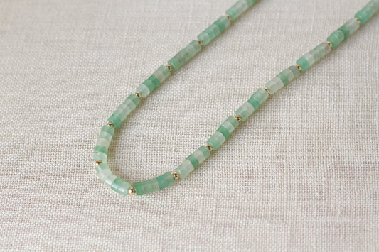 A beaded necklace made of small green stone beads separated by tiny gold spacer rings, laid on a textured off-white fabric background.