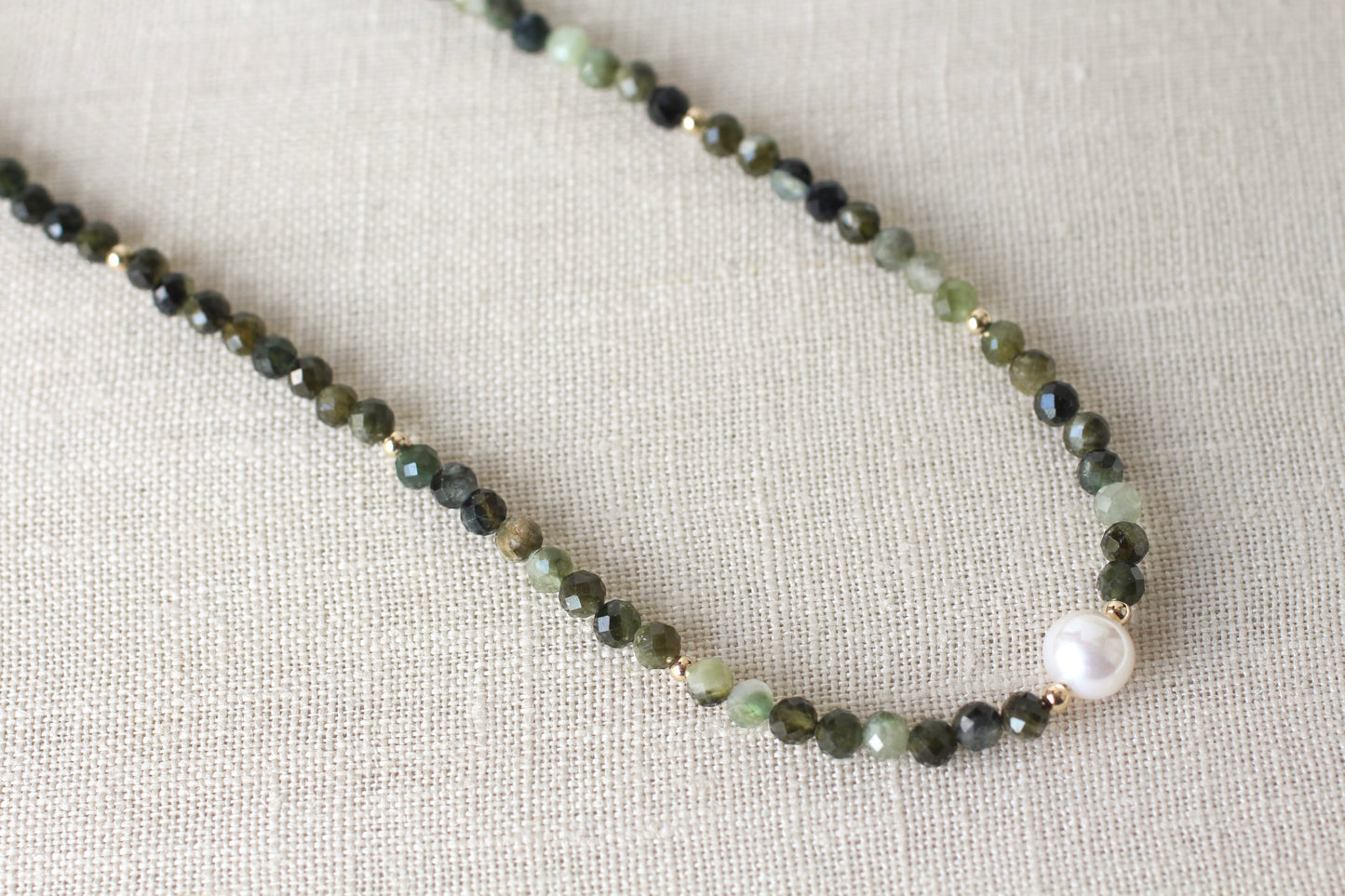 A beaded necklace featuring faceted dark green and light green beads, small gold spacer rings, and a single white pearl bead, displayed on a textured beige linen fabric background.