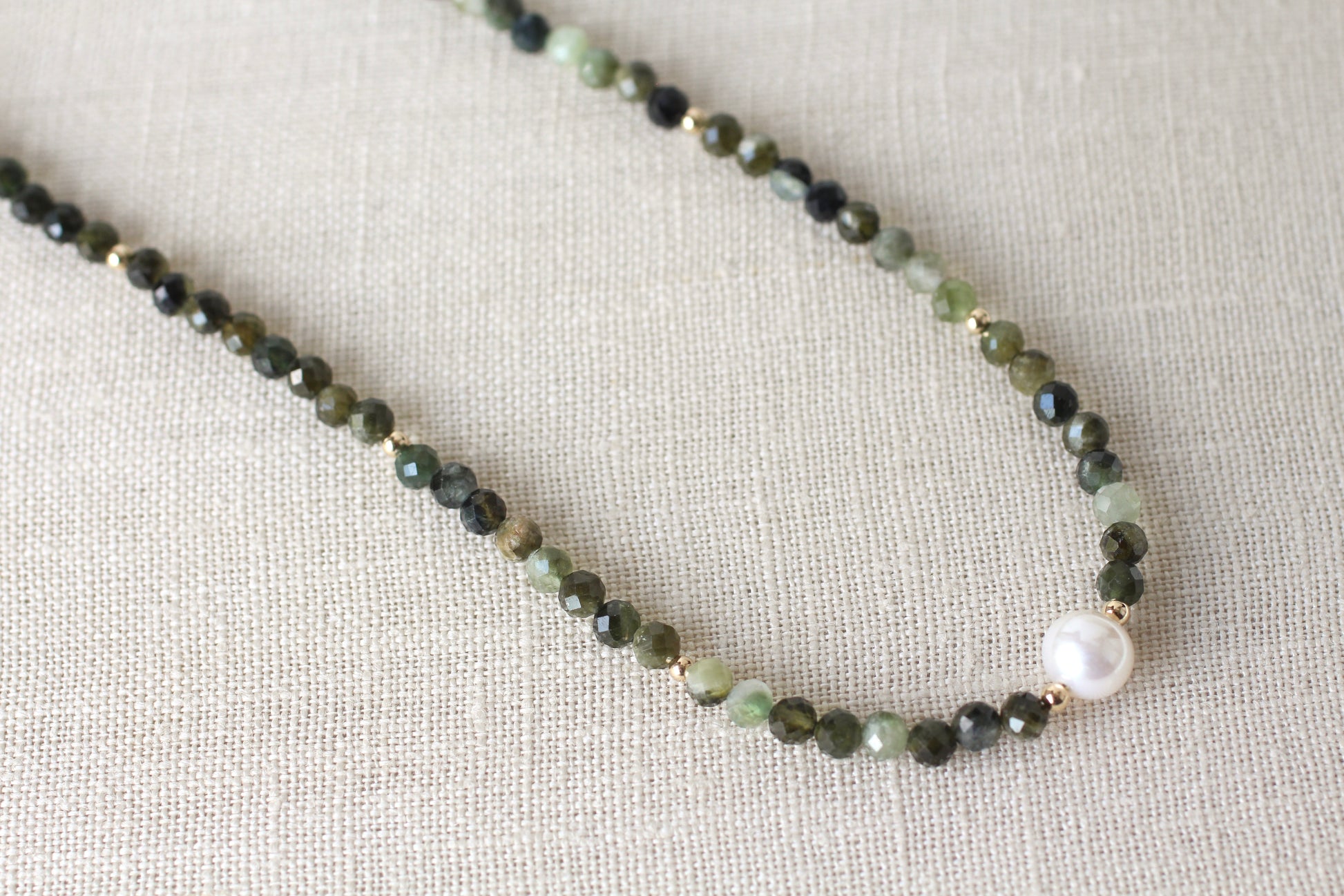 A beaded necklace featuring faceted dark green and light green beads, small gold spacer rings, and a single white pearl bead, displayed on a textured beige linen fabric background.