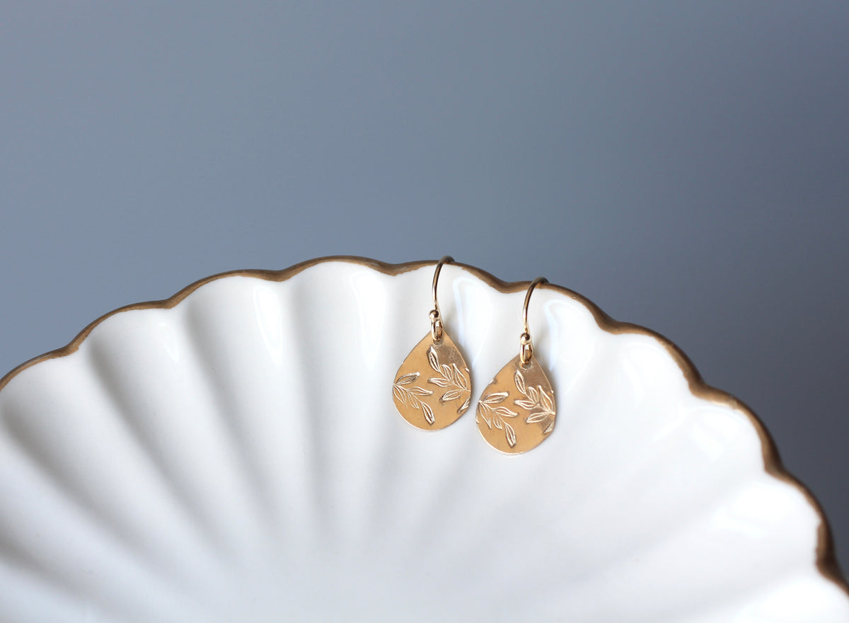A pair of gold teardrop dangle earrings with engraved leaf patterns, resting on the edge of a white scalloped ceramic dish against a solid gray background.