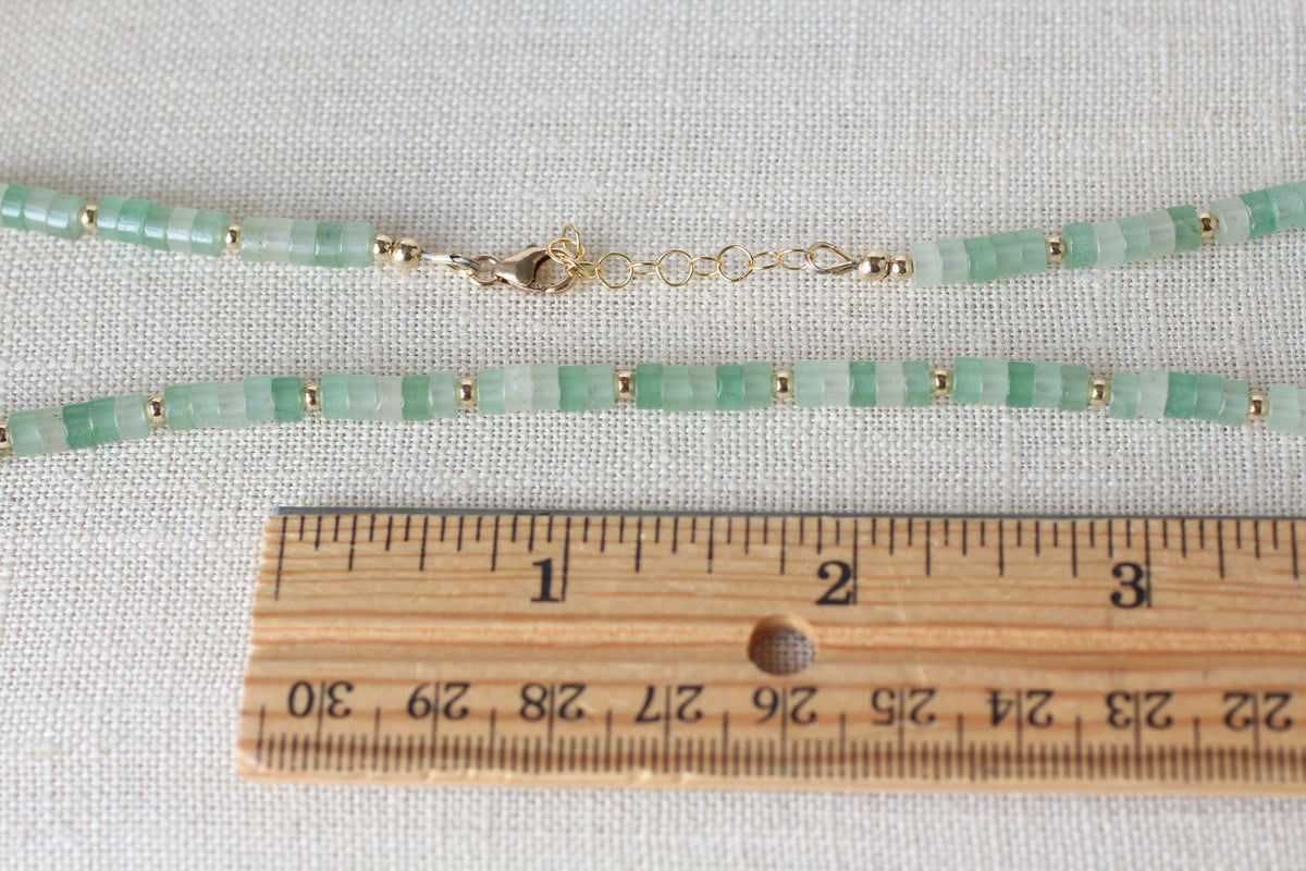 A beaded necklace made of light green and white translucent beads separated by small gold spacer beads, with a lobster clasp, displayed on a textured white fabric next to a wooden ruler for scale.