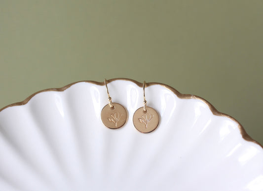 A pair of gold disc drop earrings with a delicate leaf engraving, resting on a white scalloped ceramic dish against a muted sage green background.