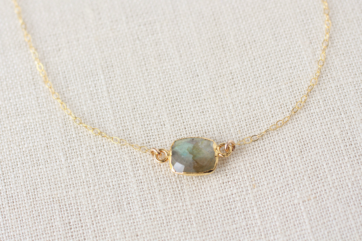 A delicate gold chain necklace featuring a small rectangular cabochon stone with a greenish-gray iridescent sheen, set in a gold bezel, displayed on a textured beige linen fabric background.