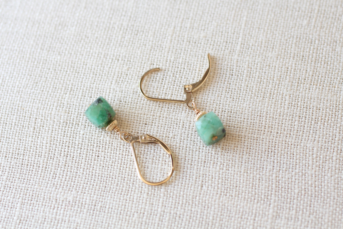 A pair of gold-toned drop earrings with small, rounded cube green beads, displayed on a textured beige linen fabric background.