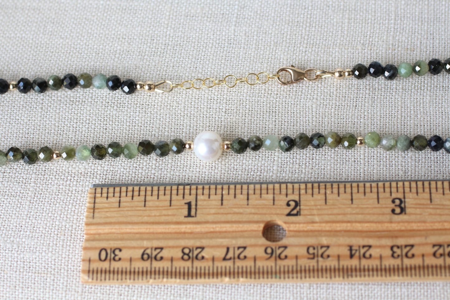 A beaded necklace with faceted dark green, light green, and black beads, a single white pearl, and gold-tone chain hardware, shown on a textured beige fabric with a wooden ruler for scale.