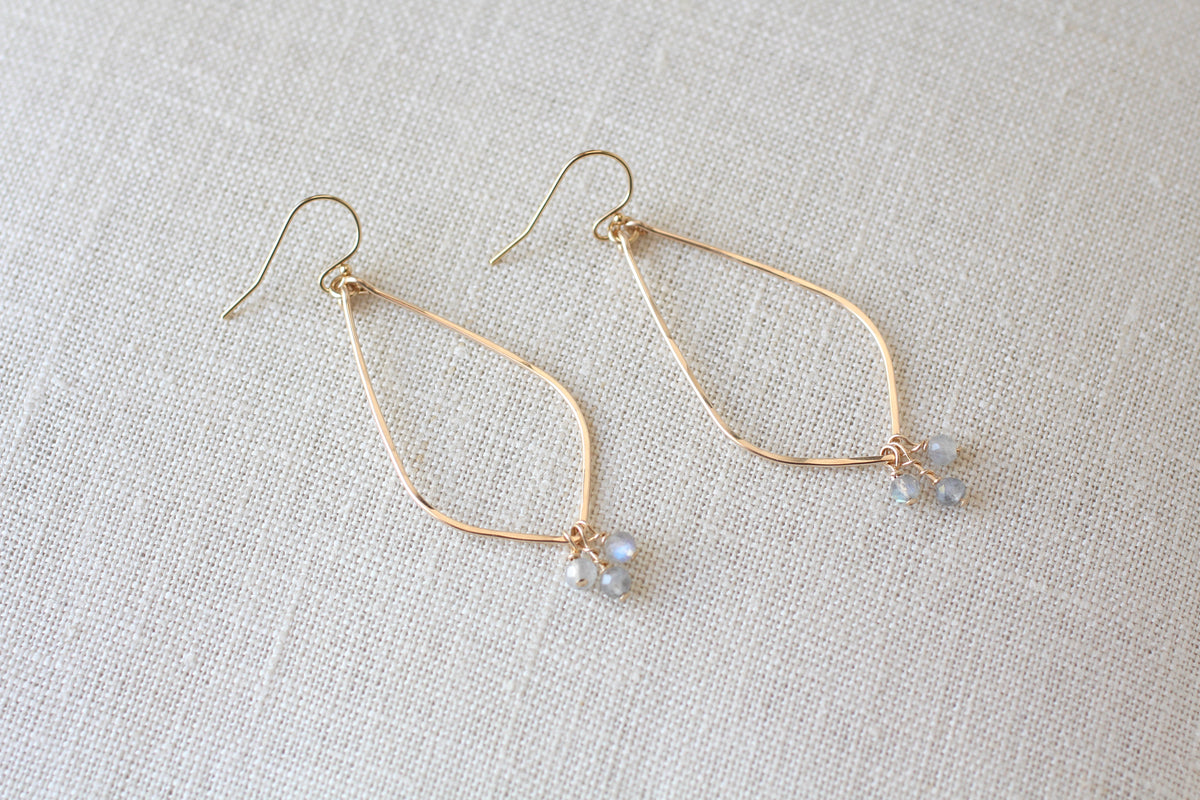 A pair of gold-tone dangle earrings with open teardrop-shaped wire frames and small gray blue bead clusters at the bottom, displayed on a textured off-white linen background.