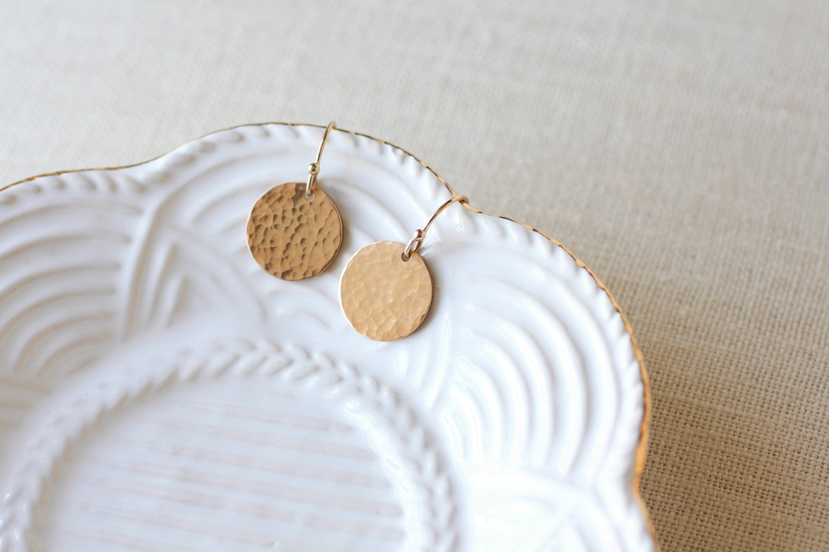 Two hammered round gold-tone dangle earrings resting on a white scalloped ceramic dish over a beige linen surface.