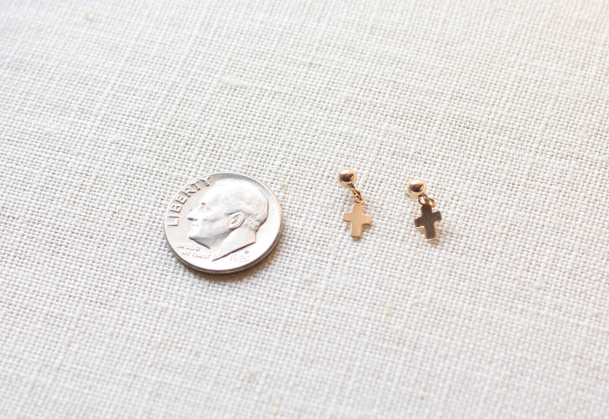Two small gold cross dangle earrings displayed next to a US dime on a textured off-white linen fabric background.