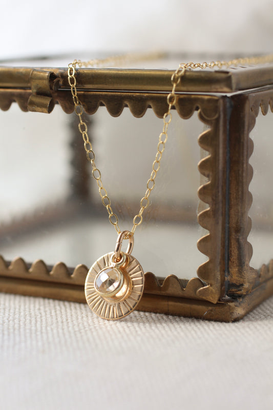 A gold necklace with a sunburst pendant and a central clear stone, displayed on a decorative bronze-toned jewelry box against a soft light background.
