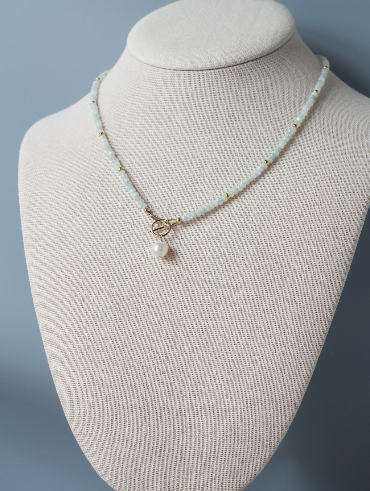 A delicate necklace displayed on a cream fabric jewelry bust. The necklace features small pale green blue beads, tiny gold spacer beads, a gold toggle clasp, and a single white pearl pendant.