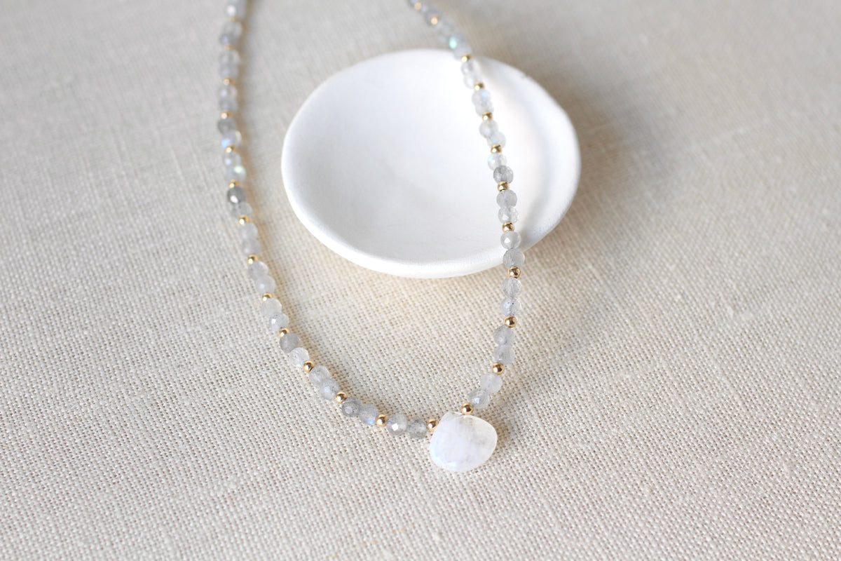 A delicate beaded necklace with pale gray and white beads, small gold spacer beads, and a round white stone pendant, displayed on a textured beige fabric next to a small white dish.