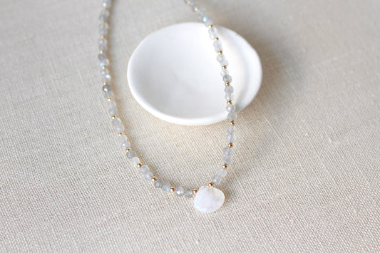 A delicate beaded necklace with pale gray and white beads, small gold spacer beads, and a round white stone pendant, displayed on a textured beige fabric next to a small white dish.