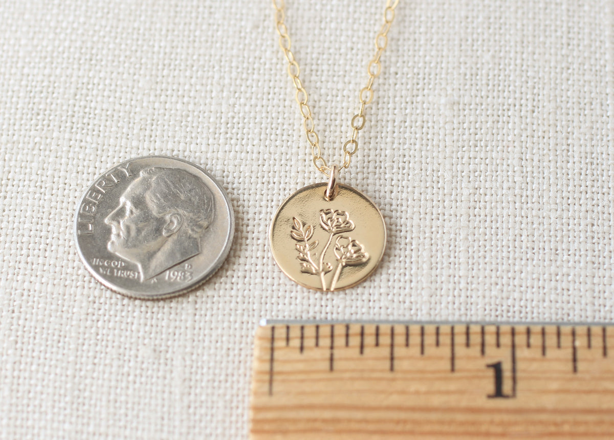 A gold coin pendant necklace with a rose engraving, displayed on a textured beige fabric next to a US dime for scale and a wooden ruler.