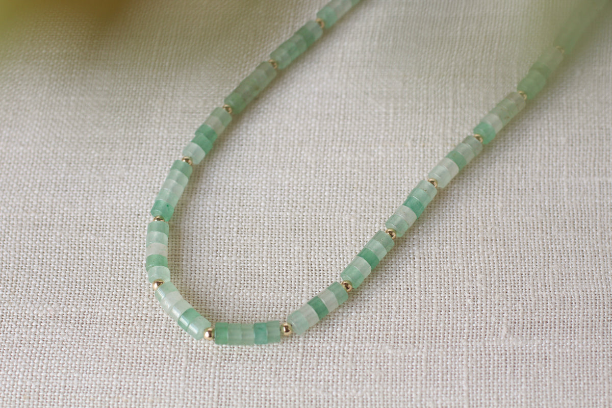 A delicate beaded necklace made of small light green stone beads separated by tiny gold spacer rings, resting on a textured beige linen surface.