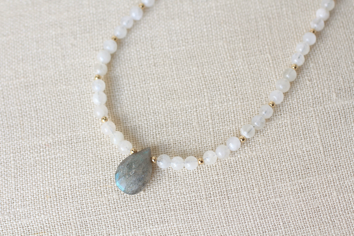 A beaded necklace featuring small white round beads separated by tiny gold spacer beads, with a central teardrop-shaped blue gray labradorite pendant, displayed on a textured beige linen fabric background.