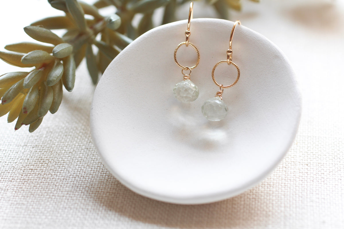 A pair of gold drop earrings with circular hoops and faceted pale green gemstones, displayed on a small white dish with a green plant in the background.