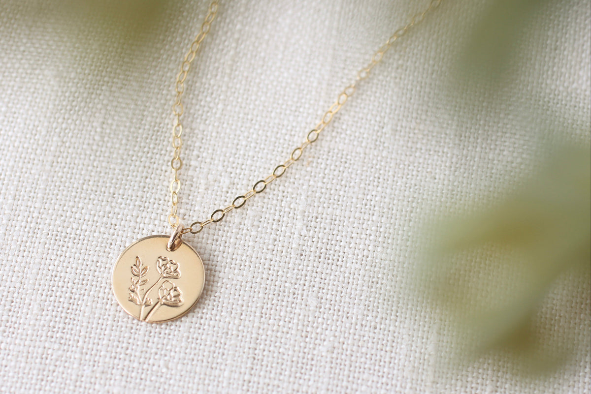A delicate gold chain necklace with a round gold disc pendant engraved with a small floral design, resting on a textured off-white fabric background with soft, blurred green foliage in the foreground.