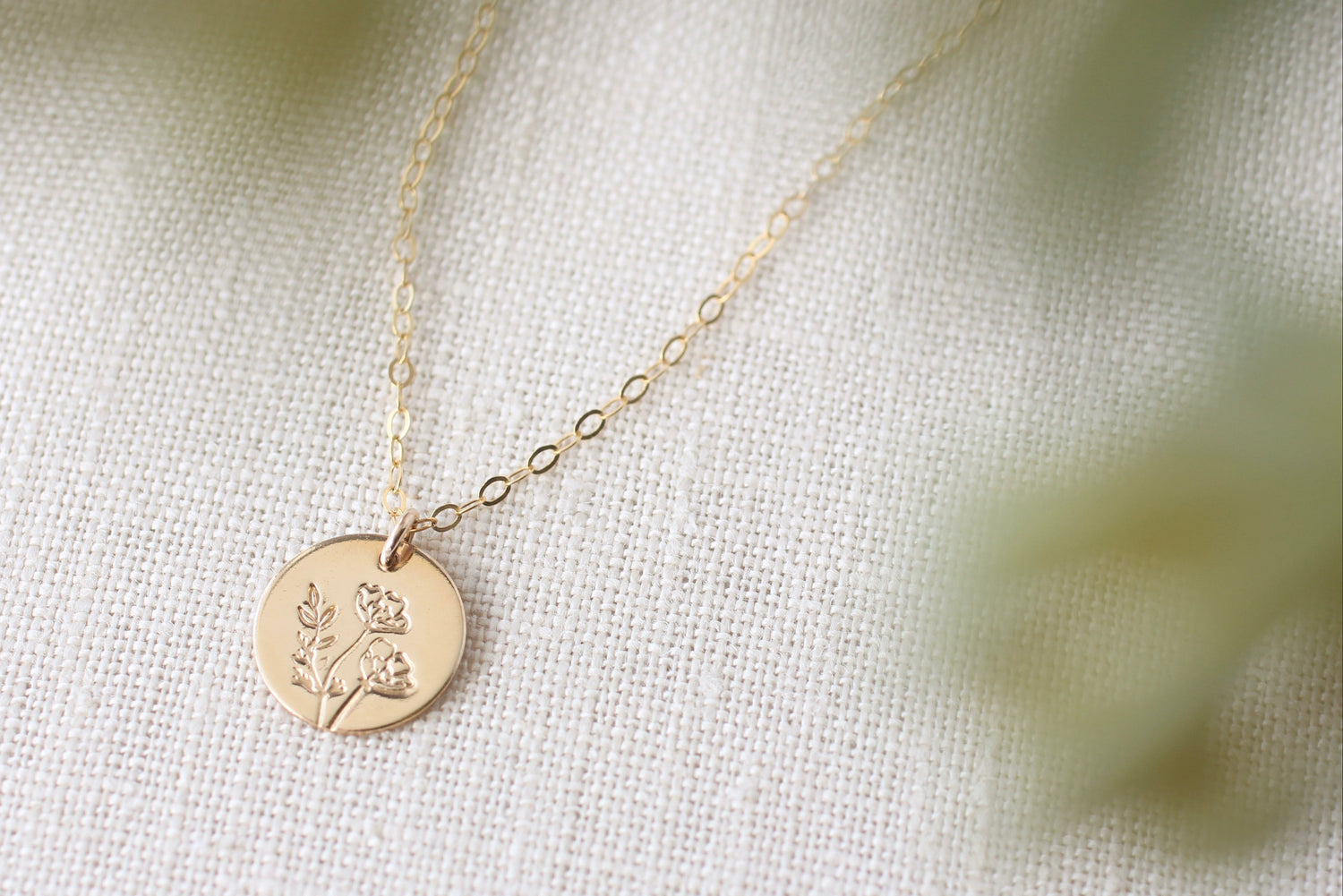 A delicate gold chain necklace with a round gold disc pendant engraved with a small floral design, resting on a textured off-white fabric background with soft, blurred green foliage in the foreground.