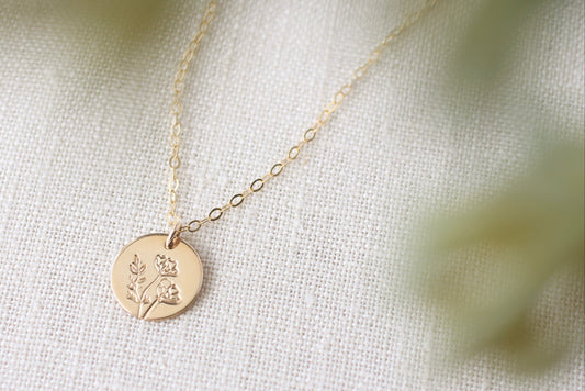 A delicate gold chain necklace with a round gold disc pendant engraved with a small floral design, resting on a textured off-white fabric background with soft, blurred green foliage in the foreground.