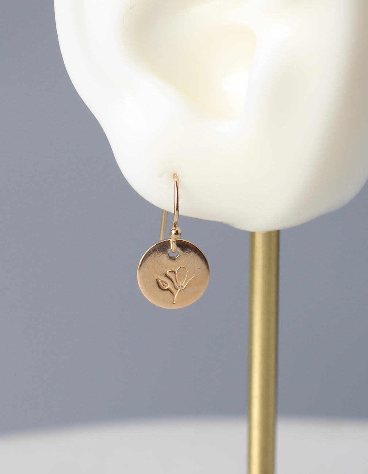 A gold disc drop earring with an engraved leaf design, displayed on a white mannequin ear against a soft gray background.